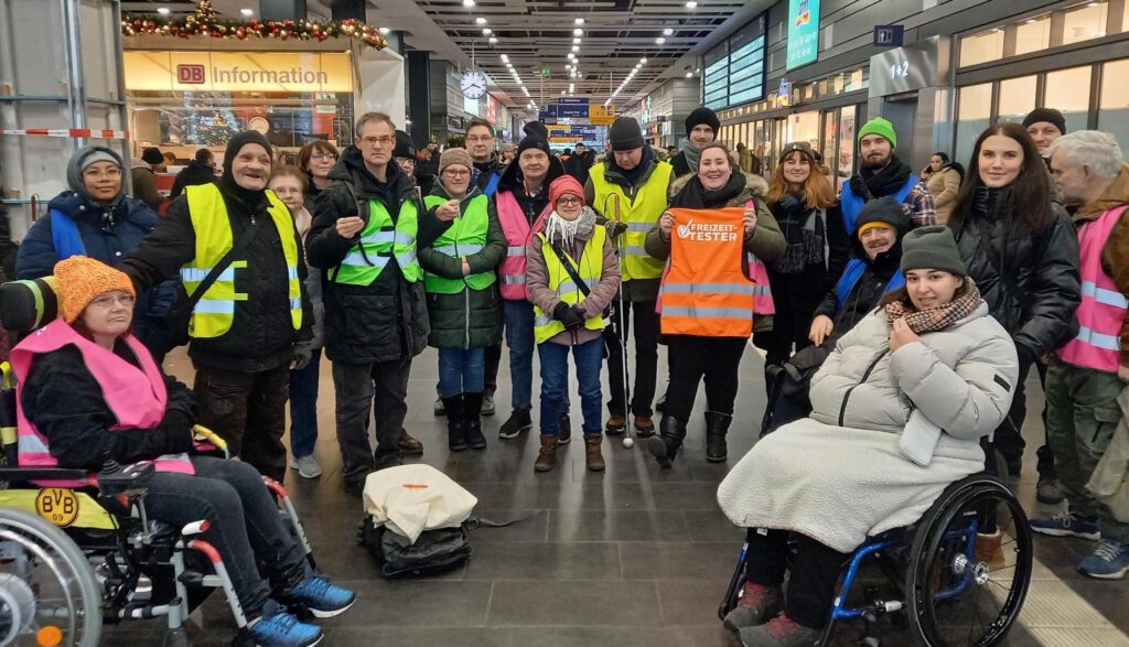 Eine Gruppe von 21 Personen mit und ohne Behinderung stehen im Halbkreis in der Halle des Essener Hbf. Sie tragen bunte Warnwesten. Es sind die Freizeit-Tester.